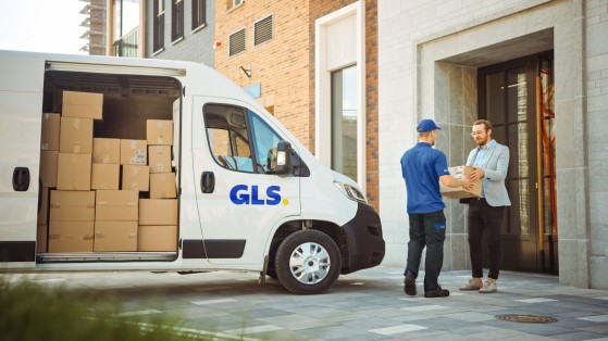 A GLS delivery van parked with its side door open and packages inside, while a GLS driver hands a parcel to a customer outside a building.