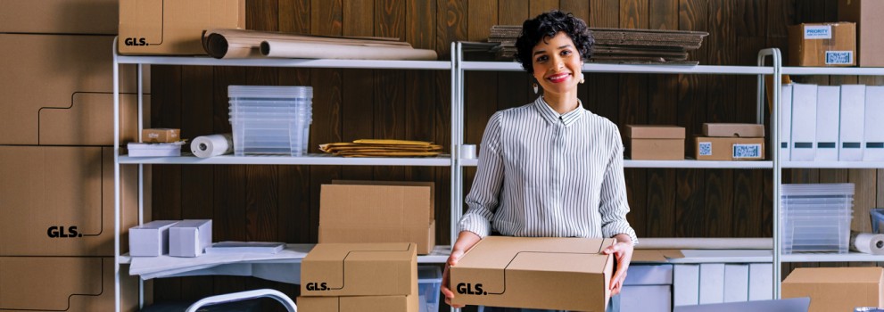 A woman standing at a table in a small warehouse, holding a cardboard shipping box. The workspace is filled with shelves of packing materials, stacked boxes, and shipping supplies.